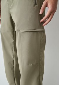 Kāju bikses olīvu zaļā audumā ar gludu faktūru, sānu kabatām un piegrieztu pieguļošumu. Izturīgs materiāls ar precīzām šuvju detaļām.