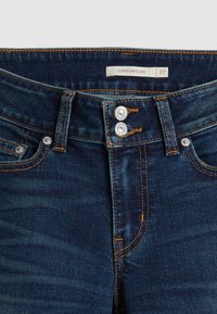 Tummansinisiä farkkuja, joissa on kaksinkertainen hopeinen nappikiinnitys, vyölenkit, etutaskut ja vyötärönsisäpuolella etiketti, jossa lukee "SUPERLOW FLARE 27".