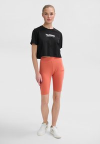 Schwarzes, kurzes T-Shirt mit "hummel sportswear"-Logo, kombiniert mit hoch taillierten korallenfarbenen Radshorts und weißen Sportschuhen.
