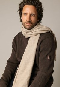 Écharpe beige en matière douce, drapée autour du cou sur un pull marron foncé côtelé. Écusson logo brun notable sur la manche gauche.