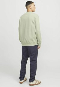 Felpa verde chiaro con polsini a coste, abbinata a pantaloni blu scuro. Modello indossa anche sneakers chiare con dettagli marroni.