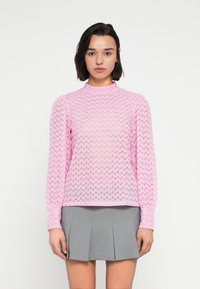 Ροζ πλεκτό πουλόβερ με ανάγλυφο σχέδιο ζικ-ζακ, ψηλό λαιμό και μακριά μανίκια, σε συνδυασμό με γκρι πλισέ φούστα.