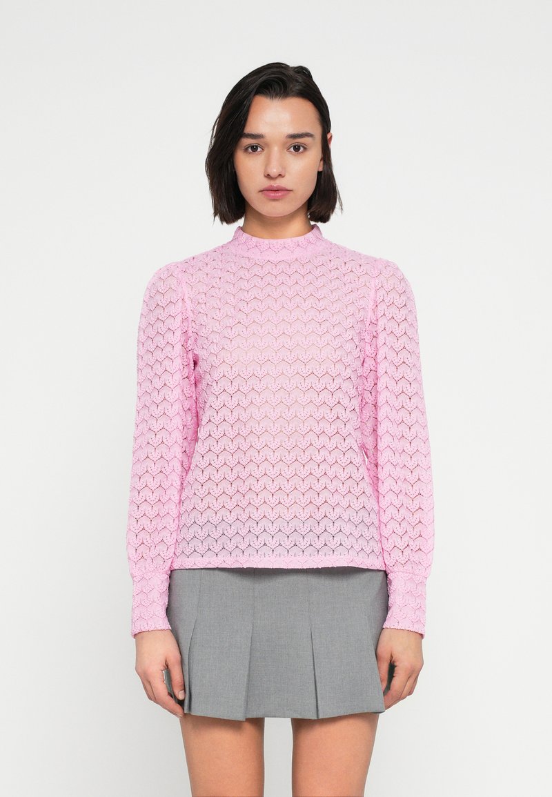 Ροζ πλεκτό πουλόβερ με ανάγλυφο σχέδιο ζικ-ζακ, ψηλό λαιμό και μακριά μανίκια, σε συνδυασμό με γκρι πλισέ φούστα.