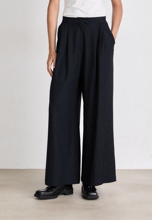 Pantaloni a gamba larga in tessuto scuro con un sottile motivo a righe, caratterizzati da una vita alta e tasche laterali, abbinati a scarpe con zeppa nere.