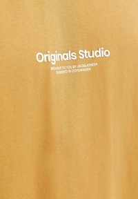 Sinepju dzeltena kokvilnas T-kreklu ar baltu tekstu: "Originals Studio" un "Pasniegts jums no JACK & JONES, izstrād�āts Kopenhāgenā."