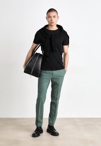 Zwart T-shirt met korte mouwen, met een gebreide trui over de schouders gelegd, gecombineerd met groene op maat gemaakte broek en zwarte veterschoenen. Draagt een zwarte tas.