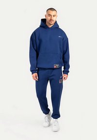 Marineblaue Kapuzenpullover und passende Jogginghosen aus weichem, dickem Material mit Logo- und Aufnäherdetails, kombiniert mit weißen Turnschuhen.