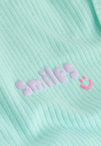 Tissu côtelé vert clair avec le mot « smiles » en lettres violettes pastel en relief et un petit visage souriant rose à côté.