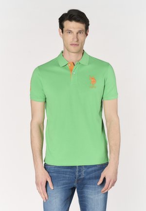 Uomo che indossa una polo verde a maniche corte con logo e dettagli arancioni, abbinata a jeans blu, in piedi davanti a uno sfondo bianco.