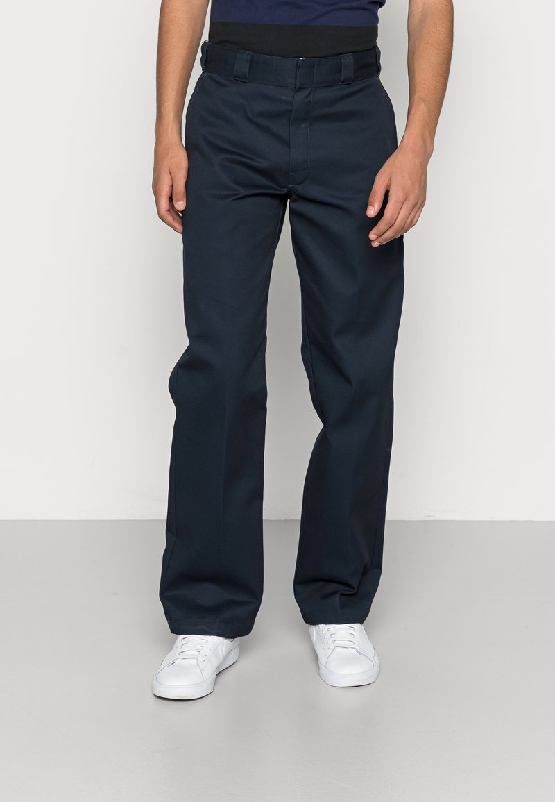 Homme portant un pantalon droit bleu marine et des baskets blanches, debout contre un mur et un sol gris clair.