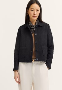 Schwarze, texturierte Jacke mit einem cropped Design, zwei frontalen Taschen und blauen Knöpfen. Wird über einem braunen Oberteil mit einem gestreiften Schal getragen.