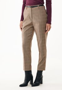 Pantaloni a quadri marroni e beige con dettagli arancioni, caratterizzati da una vestibilità su misura, pieghe frontali e una cintura nera. Indossati con stivali neri con tacco.