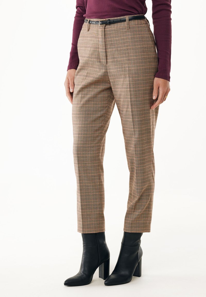 Pantalones de cuadros en marrón y beige con acentos naranjas, que cuentan con un corte ajustado, pliegues en la parte delantera y un cinturón negro. Combinados con botas de tacón negras.