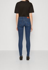 Persona vestida con jeans ajustados de color azul oscuro y zapatos negros, de pie frente a una pared blanca lisa, con una blusa negra de manga corta.