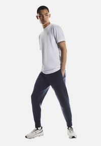 Hellgraues Sport-T-Shirt mit kurzen Ärmeln und Logo, kombiniert mit marineblauen Jogginghosen und weißen Turnschuhen mit schwarzen Akzenten.