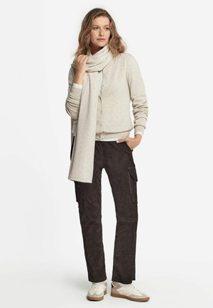 Femme portant un cardigan beige et une écharpe assortie, un pantalon cargo foncé et des baskets blanches, debout avec une main dans la poche.