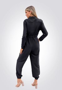 Combinaison noire à manches longues, taille élastique et chevilles resserrées. Dotée d'un col et d'un tissu lisse. Associée à des talons beiges.