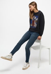 Sudadera negra con un diseño gráfico colorido, combinada con unos jeans ajustados azules y zapatillas blancas. Sentado en una silla blanca sencilla.