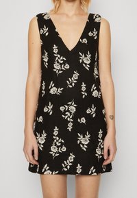 Vestido negro con un escote en V profundo y bordados florales en blanco. Diseño sin mangas, silueta ajustada y dobladillo corto. Tejido texturizado.