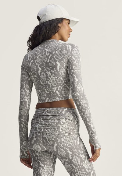 Mujer con gorra blanca y conjunto a juego de top de manga larga y leggings gris con estampado de piel de serpiente, de espaldas.