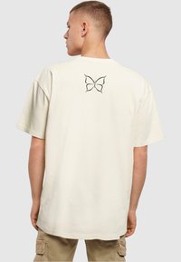 Lys beige oversized t-shirt lavet af bomuld, med en sommerfuglegrafik trykt i sort på den øverste del af ryggen. Korte ærmer og rund halsudskæring.