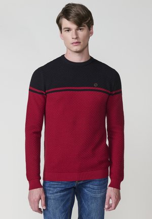 Suéter texturizado rojo y negro con cuello redondo, que presenta rayas horizontales y un pequeño emblema en el pecho. Combinado con jeans azules.