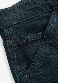 Donkerdenim jeans met een gestructureerde afwerking, voorzien van oranje stiksels en een "SUPPLY Co." label op de zak. Inclusief details van messing klinknagels.