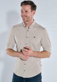 Lächelnder Mann mit kurzen Haaren, der ein hellbeiges, knöpfbares Hemd mit aufgerollten Ärmeln und blaue Jeans trägt, vor einem schlichten Hintergrund stehend.