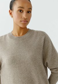 Beige gestrickter Pullover mit rundem Halsausschnitt und Rippdetails an den Ärmeln, aus weichem Material gefertigt und leicht locker geschnitten.