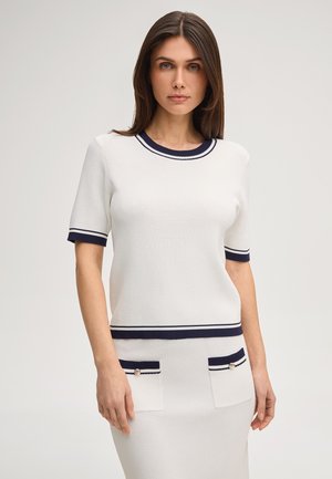 Frau trägt ein weißes Strickoberteil und einen Rock mit marineblauem Rand und taschen mit Knöpfen, steht vor einem schlichten Hintergrund.