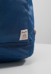 Zaino in tessuto blu con superficie testurizzata e dettagli marroni. Presenta un'etichetta bianca con il logo "Doughnut Outdoor Adventure".