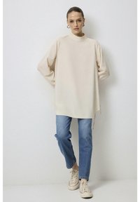 Haut beige à manches longues avec un col haute, coupe décontractée, fentes latérales, associé à un jeans bleu clair et des baskets beiges. Design simple.