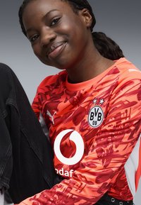 Oranssi kuviollinen urheilu asunto mustilla yksityiskohdilla, jossa on pyöreä logo ja hiusyksityiskohtia. Mallilla on päällä mustat housut, istuu neutraalilla ilmeellä.