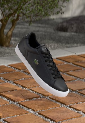 Zapatilla de cuero negra con cordones negros, suela blanca y logo verde de Lacoste. Textura suave con un diseño minimalista.