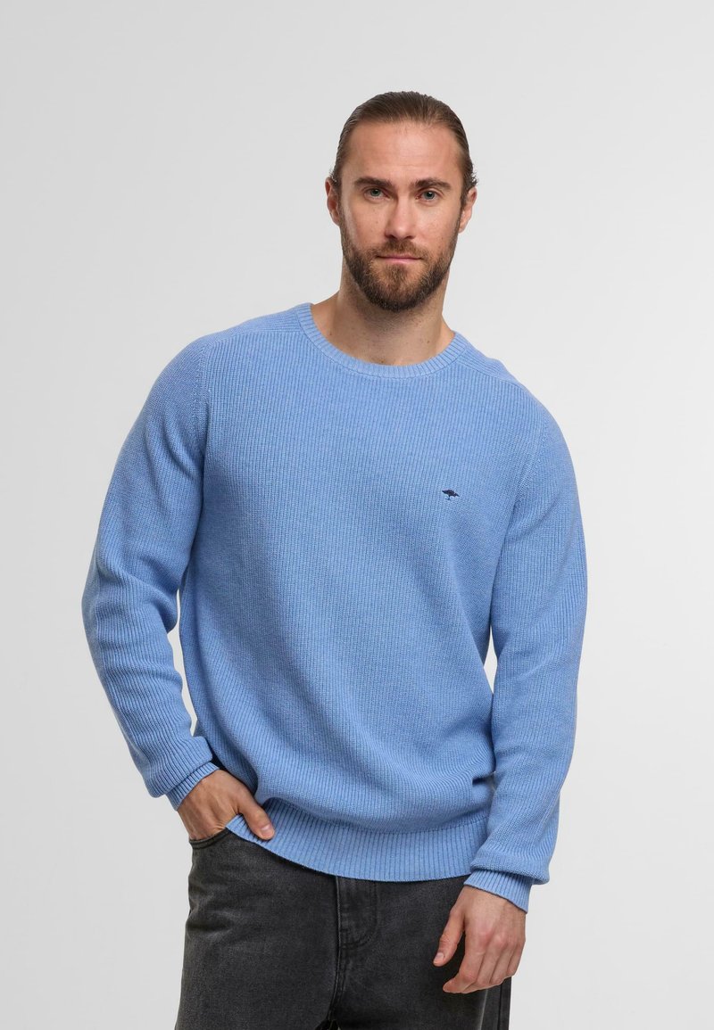 Mann mit Bart und zurückgebundenem Haar, der einen hellblauen Strickpullover und schwarze Jeans trägt, posiert mit einer Hand in der Tasche vor einem weißen Hintergrund.