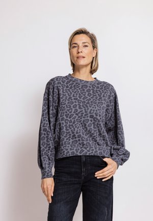 Woman with short blonde hair wearing a gray leopard print sweatshirt and black jeans standing against a plain white background.