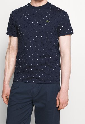 Mężczyzna w granatowej koszulce z krótkim rękawem w białe kropki i małe zielone logo krokodyla, w połączeniu z ciemnogranatowymi spodniami.