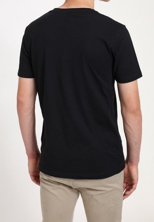 Mann trägt einfaches schwarzes T-Shirt mit kurzen Ärmeln und beige Hose, steht mit dem Rücken zur Kamera vor einem weißen Hintergrund.