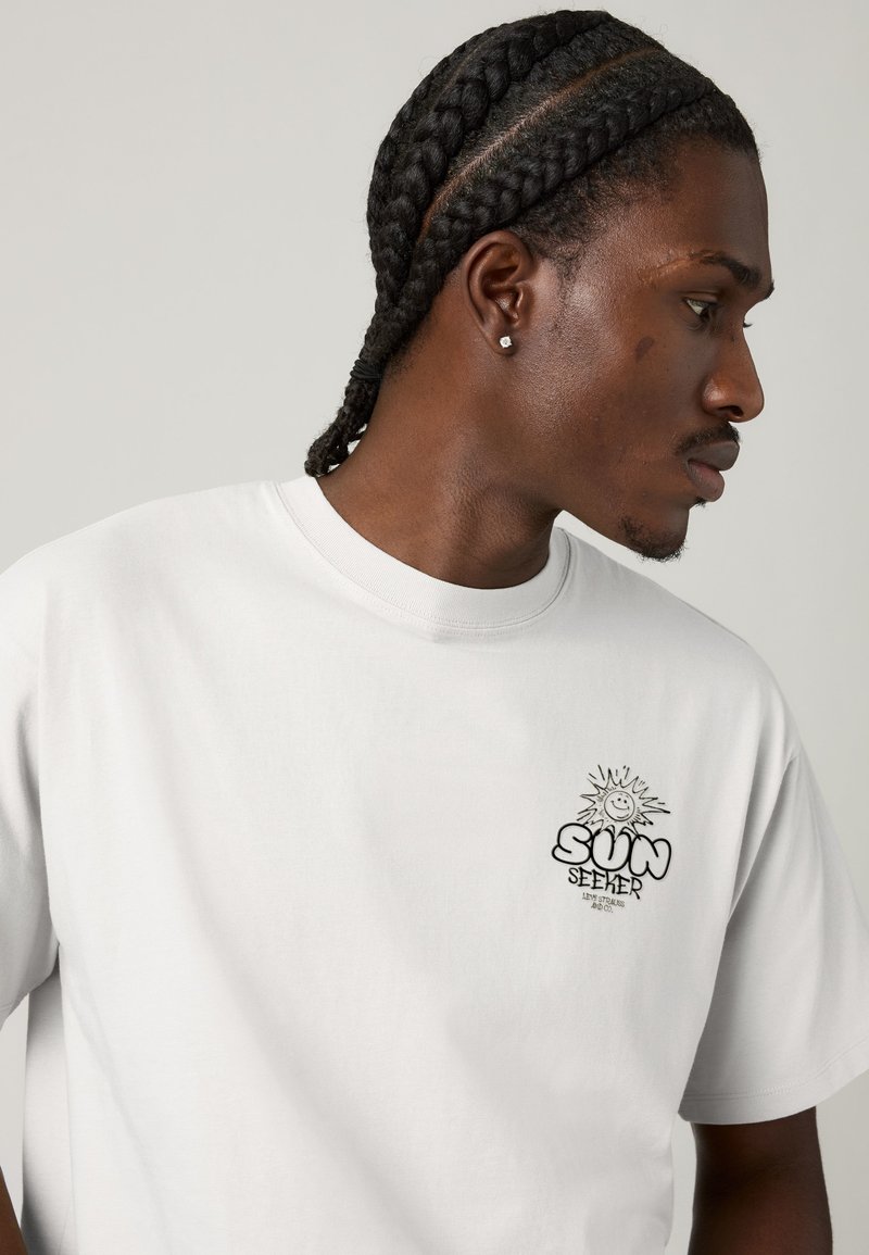 Ung mand med flettet hår bærer en hvid T-shirt med en lille solgrafik og teksten "SUN SEEKER" på brystet, kigger til siden.