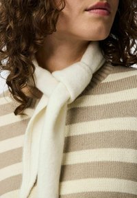 Kvinde med krøllet hår iført en beige og hvidstribet sweater og et blødt, cremet tørklæde bundet om halsen.