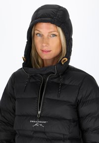 Svart pufferjacka med hög huva, med dragkedjestängning fram och dragsnoddar. Logotypen syns på bröstet. Slät, quiltad textur.