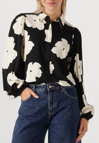Zwarte blouse met groot wit bloemenpatroon, knoopsluiting aan de voorkant en pofmouwen, gecombineerd met hooggesneden blauwe spijkerbroek.