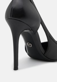 Chaussure à talon haut en cuir noir au design épuré, avec des découpes courbées et un détail en métal portant le logo sur le talon. Texture lisse, bout pointu.