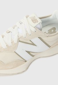 Baskets New Balance beige et blanches avec lacets blancs, dessus en daim et mesh, et logo « N » de la marque sur le côté.