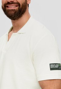 Weißes Polohemd mit strukturiertem Muster, kurzen Ärmeln, einem Kragen und einem schwarzen Etikett am Ärmel mit weißem Text.