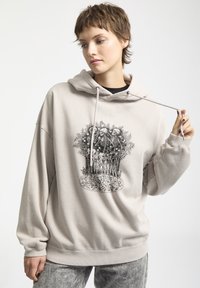 Beżowy, oversize'owy hoodie z kapturem regulowanym sznurkiem. Posiada czarny nadruk graficzny z czaszkami otoczonymi liśćmi. Miękka tkanina, luźny krój.