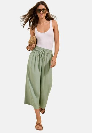 Kobieta w zielonych szerokich spodniach, białym topie na ramiączkach, brązowych sandałach i dużych okularach przeciwsłonecznych, trzyma plecioną kopertówkę i idzie do przodu.