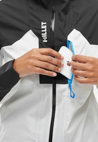 Hände, die ein weißes Smartphone in eine mit einem Reißverschluss versehene Brusttasche einer schwarz-weißen Millet-Jacke mit blauem Zipper ziehen.