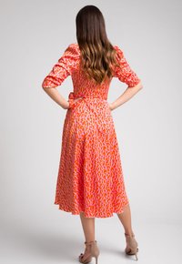 Czerwona sukienka z wzorzystym wzorem zawierającym pomarańczowe kształty, z rękawami o średniej długości, wcięciem w talii i zwiewną spódnicą. Widok z tyłu; widoczne sandały na paski.