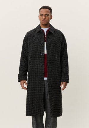 Mann trägt langen, dunkel karierten Mantel über rotem und weißem Pullover und grauer Fensterpaneel-Hose, steht vor schlichtem hellem Hintergrund.